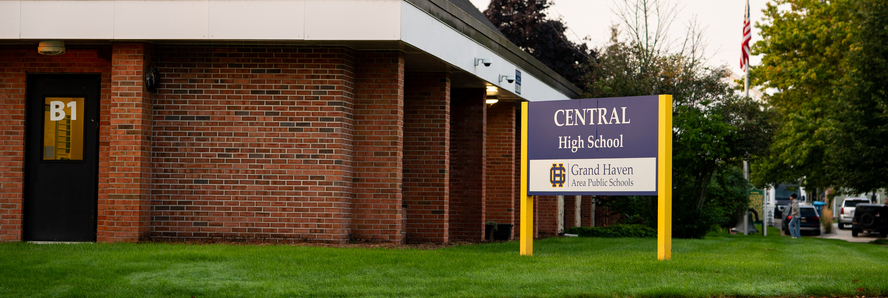 A brick school building with a peaked roof and a sign in front that reads, “Central High School, Grand Haven Area Public Schools.” The sign has a purple background with gold posts and the district’s logo. A doorway labeled “B1” is visible on the left.