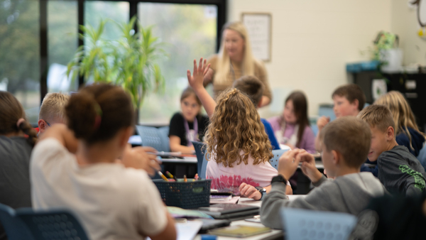 A girl raising her hand in the middle of a classroom filled with students and her teacher speaking in the background. The table the young girl is sitting at includes a basket of supplies like pencils and markers.