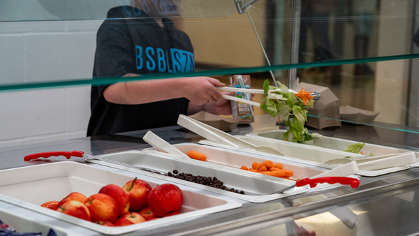 A young boy using tongs to pick up salad at the salad bar in the lunch line at school.