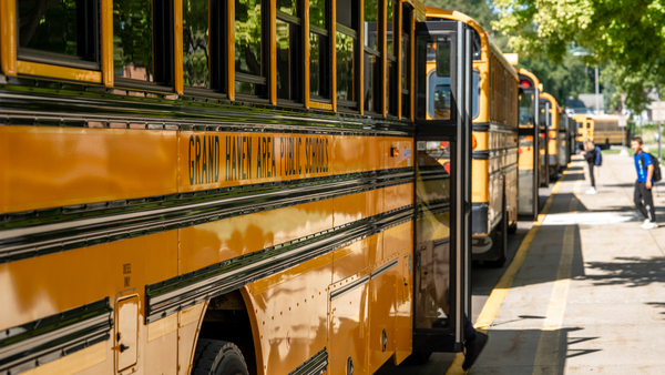 A line of Grand Haven Area Public Schools school buses parked along the sidewalk as several students begin to board them.