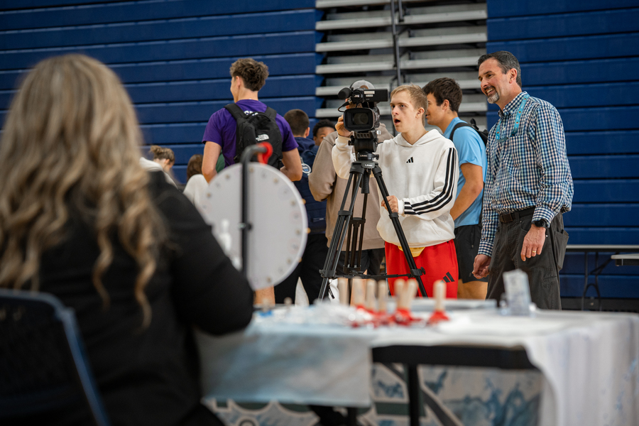 A student operating a video camera during a school event with a staff member looking on and smiling.