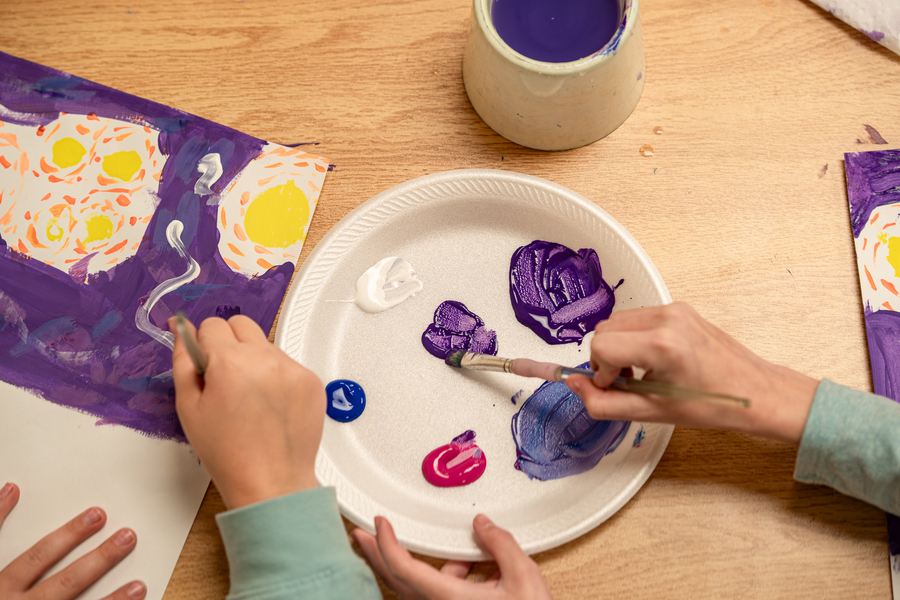 A downward perspective of two students painting purple, yellow, and orange pictures at a table in art class.