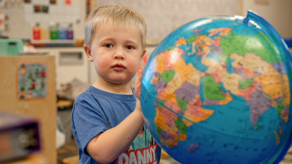 A young child looking at the camera while playing with a globe in his preschool classroom.