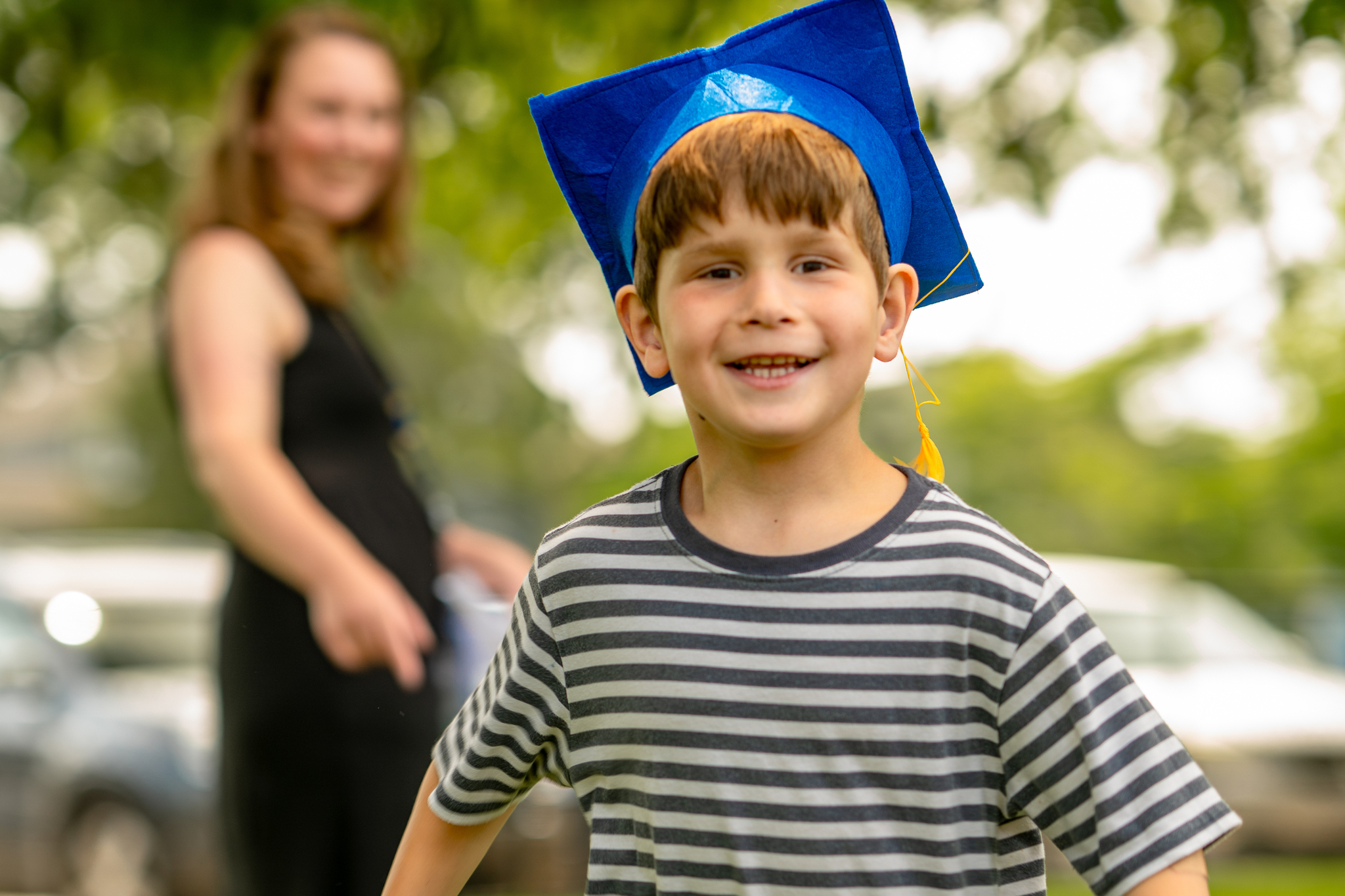 A young preschool student smiling with a grad cap on while his teacher smiles in the background.