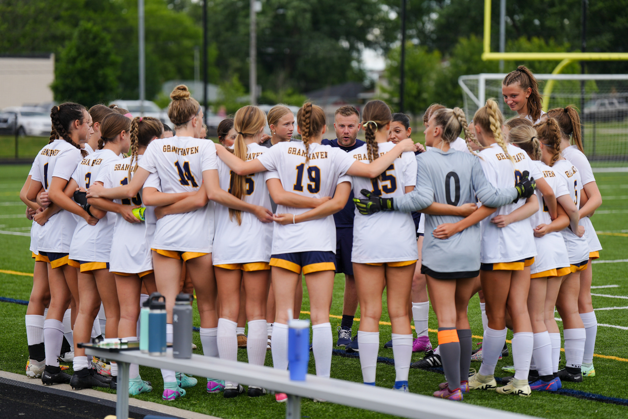 the Grand Haven girls soccer team huddled together with the head coach at the center speaking to them.