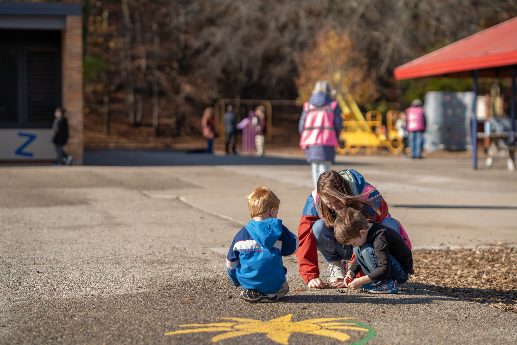 Two students playing on the pavement of a playground with a lunchsupervisor