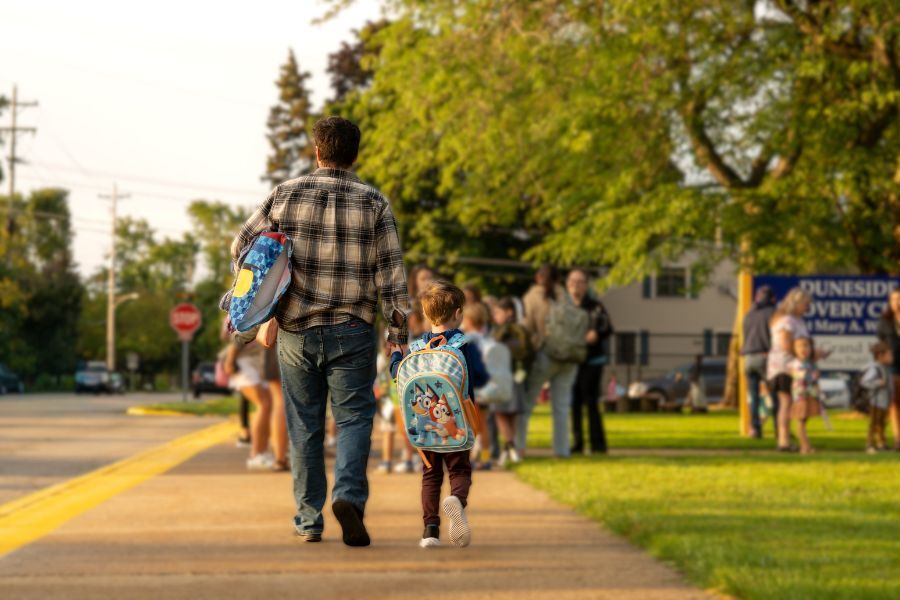 A dad walking a young boy down the sidewalk to school, pictured from behind.